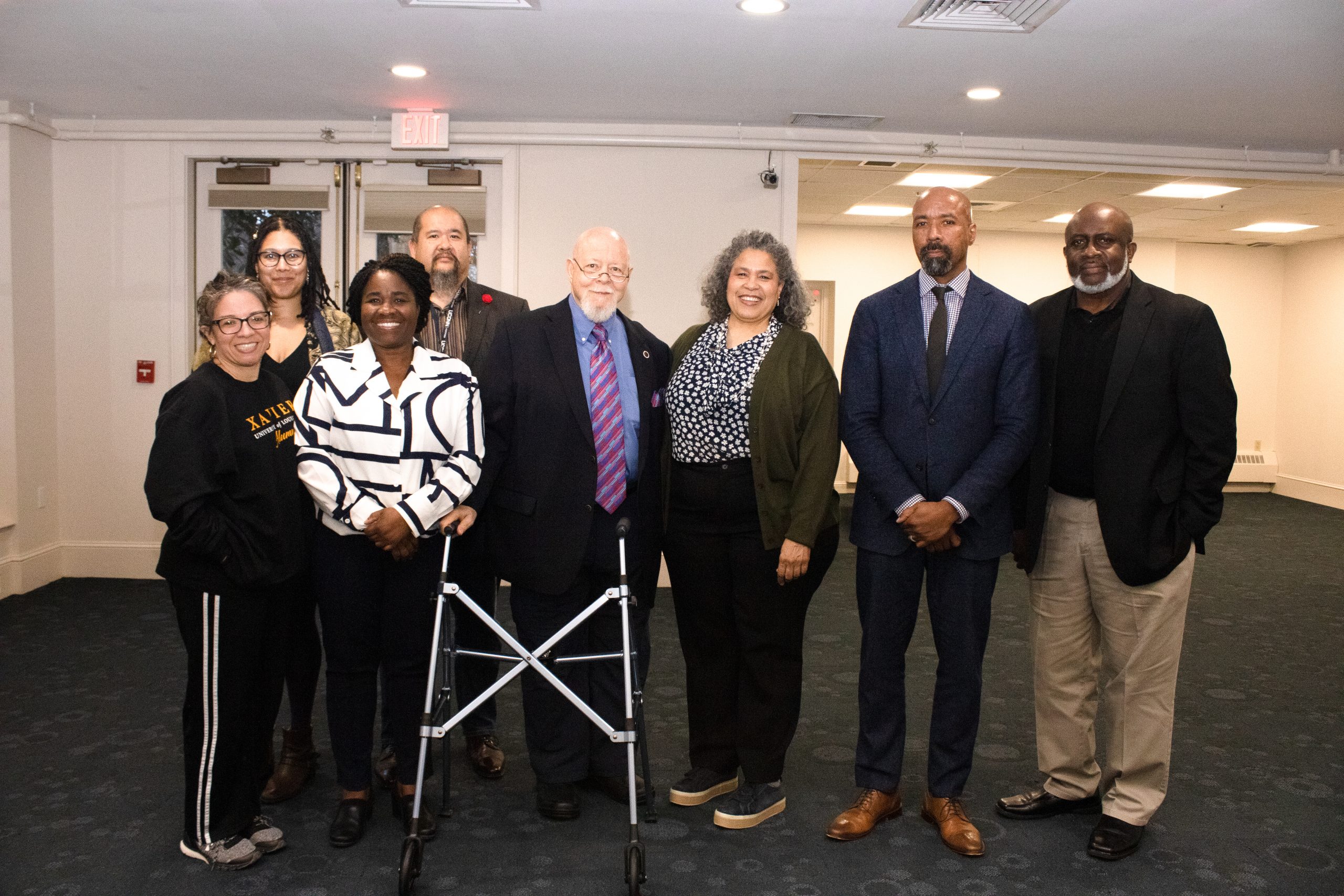 ASI Staff and Dr. Molette at the Mollette Dinner 2023: A group of eight individuals stands in a line indoors for a photo, with one of them using a mobility aid, a walker. They are dressed in a mixture of formal and casual attire. The background features an exit sign above a door on the left.